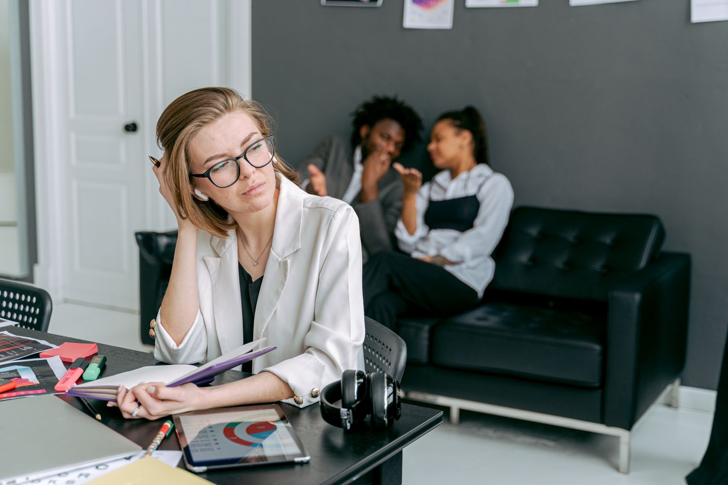 Trabalhadora visivelmente estressada em sua mesa enquanto colegas fazem fofoca ao fundo no escritório