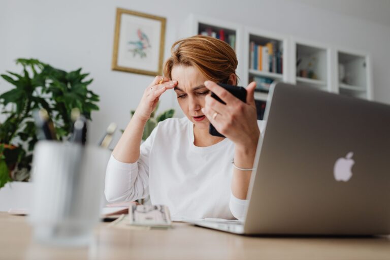 Mulher preocupada segurando o celular em mesa de trabalho com notebook