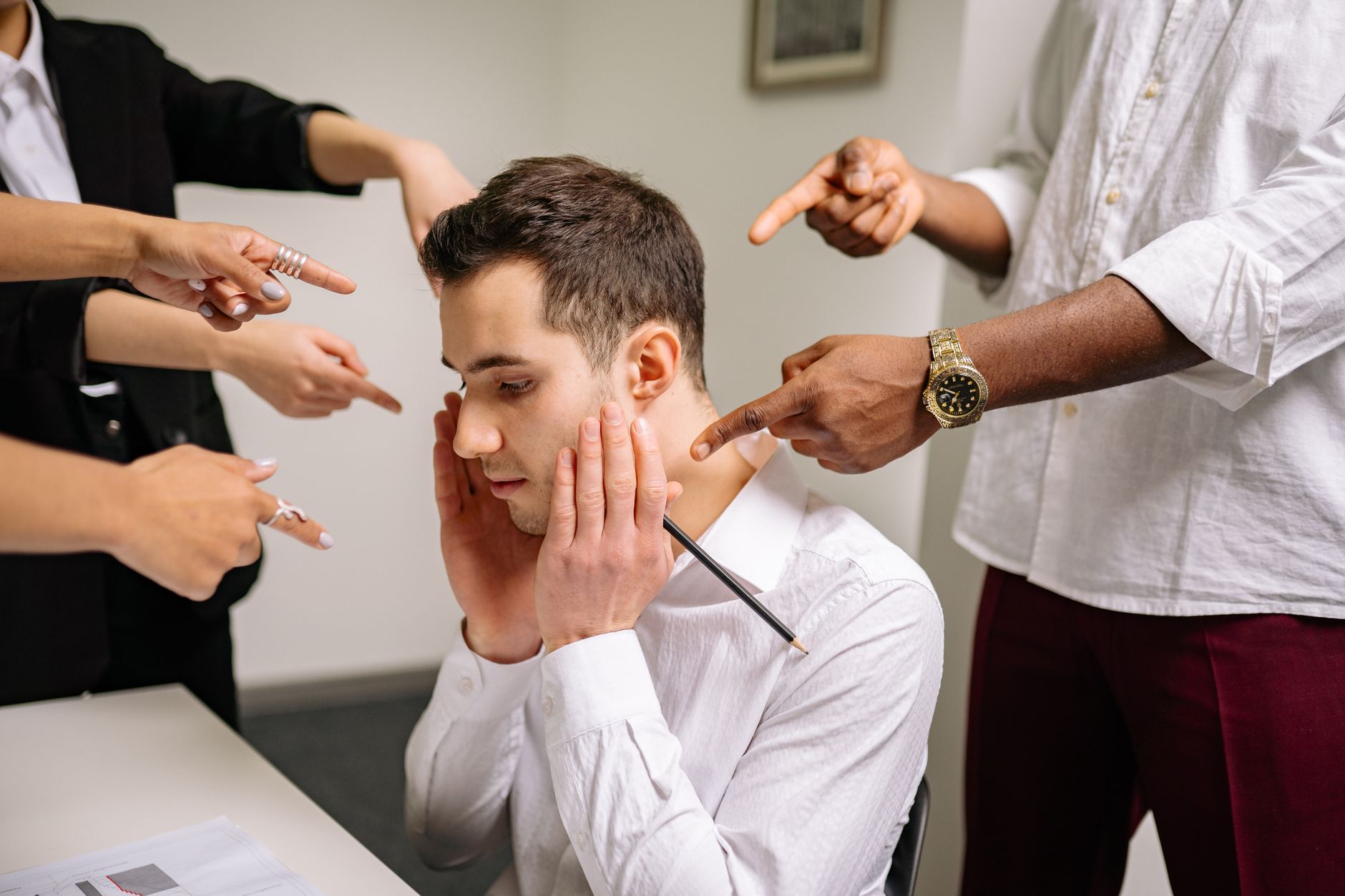 Homem estressado em escritório cercado por dedos apontados, simbolizando pressão e humilhação no trabalho
