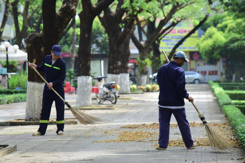 Dois trabalhadores varrendo calçada arborizada