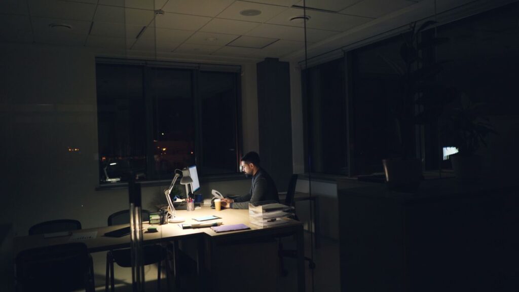 Trabalhador fazendo horas extras à noite em escritório iluminado por luminária de mesa